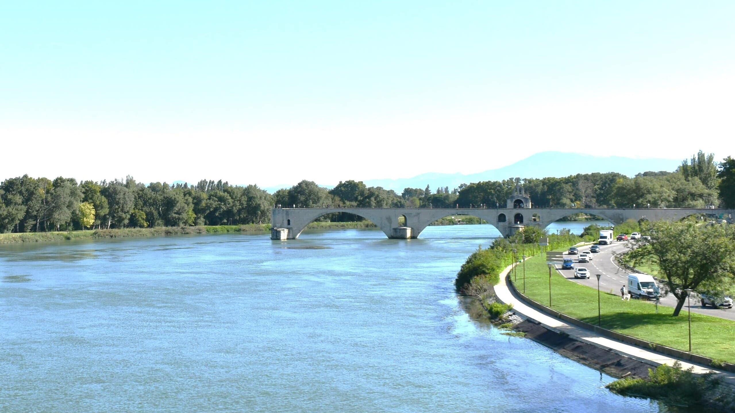 Stone bridge over a wide river with trees and a road with cars along the riverbank on a clear day, one of the 5x Best things to Do in Avignon for stunning views and memorable walks.