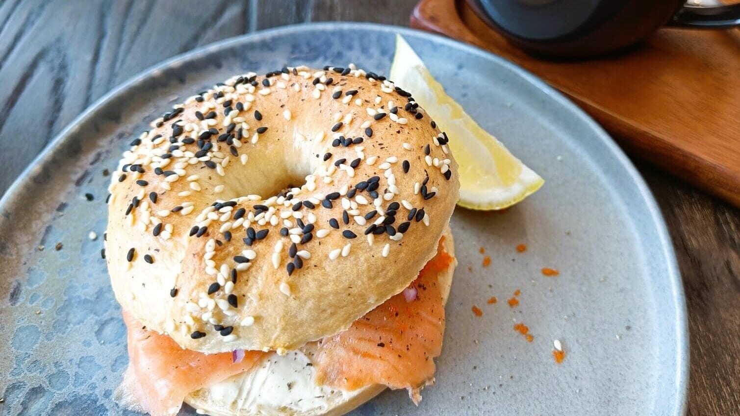 Bagel with cream cheese and salmon on a plate with a lemon wedge, accompanied by latte art coffee beside it on a wooden tray. Discover this delight at one of the best coffee shops in New York City, where every bite and sip promises an unforgettable experience.