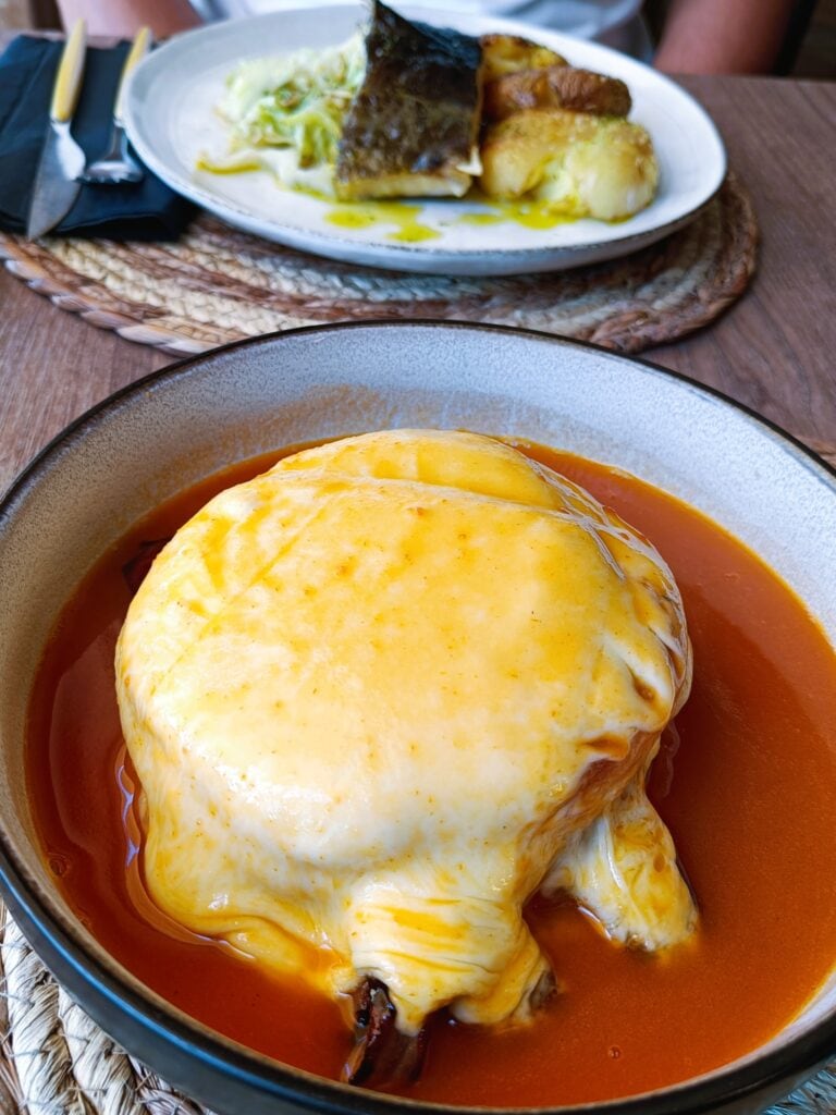 A bowl of food covered in melted cheese and sauce, reminiscent of a comforting meal you might enjoy during one day in Porto, with another plated meal blurred in the background.