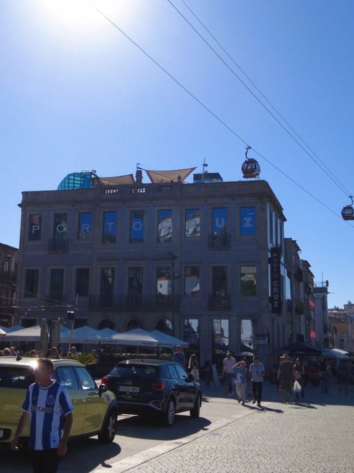 People walk by a building with "PORTO CRUZ" signs, cars parked, and cable cars above under a bright blue sky—a vibrant scene that captures the essence of one day in Porto.