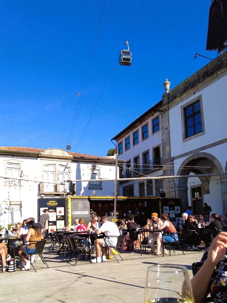 People sit at outdoor tables in a sunny square, enjoying the vibrant atmosphere of one day in Porto; cable cars travel overhead against a clear blue sky.