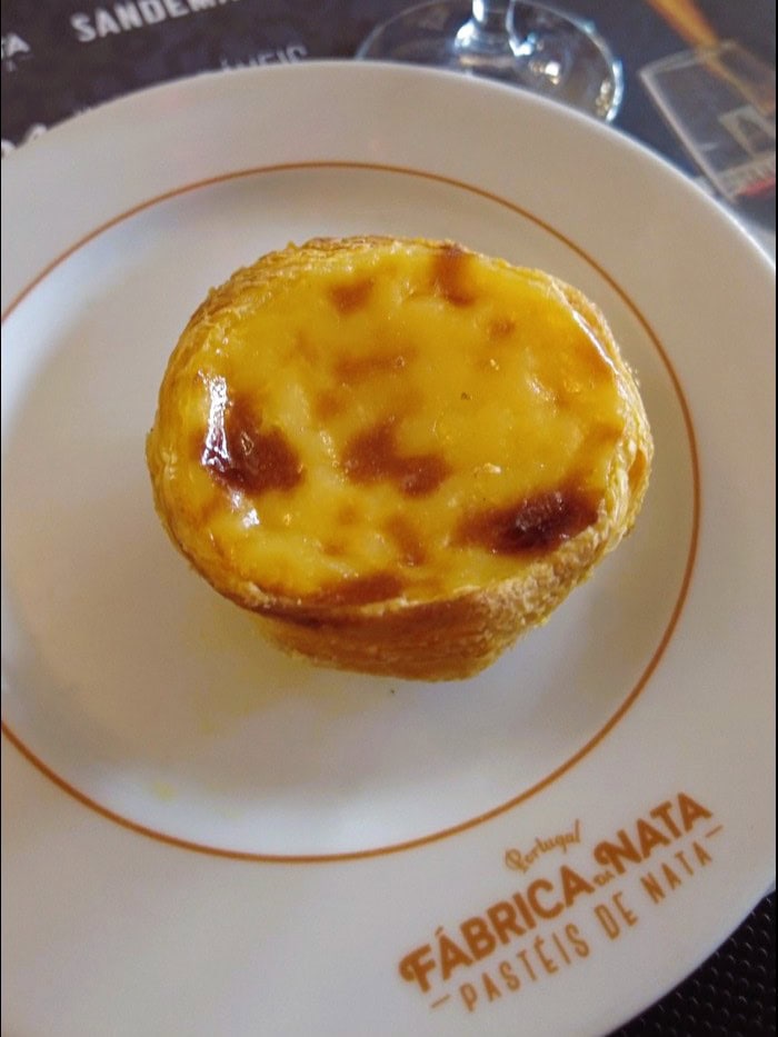 A pastel de nata sits on a white plate with "Fábrica da Nata" branding, the perfect treat to enjoy during one day in Porto.