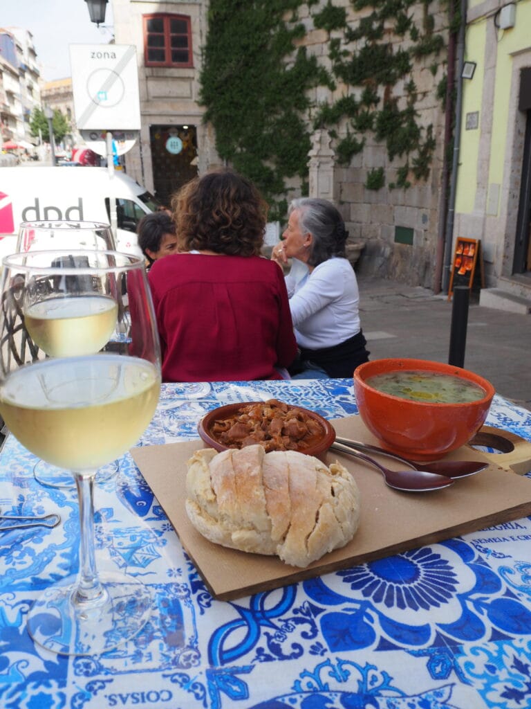 Two glasses of white wine, bread, stew, and soup on a blue tablecloth capture the essence of one day in Porto, with people talking in the background.
