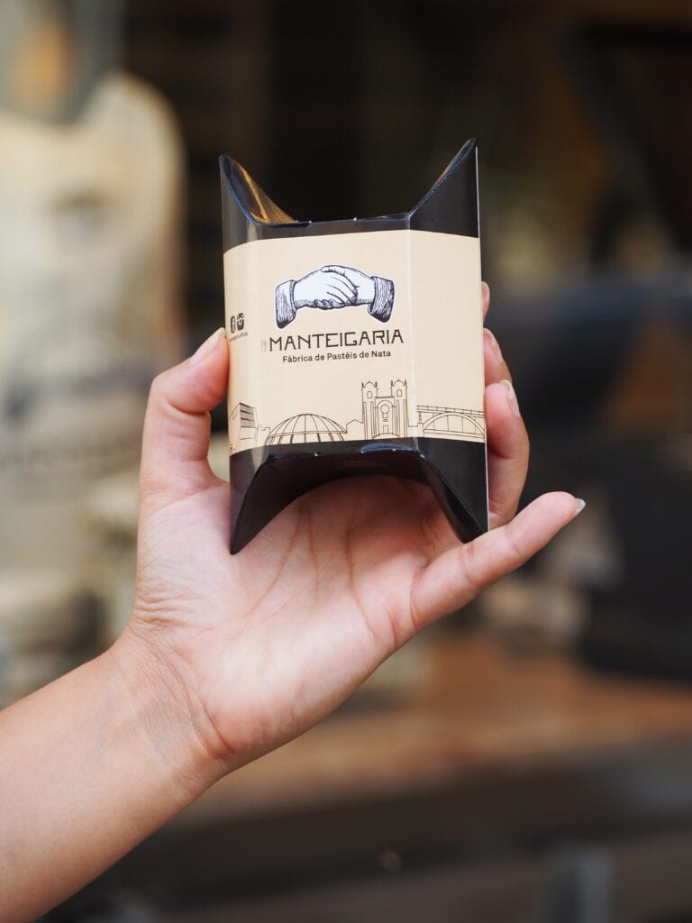 A hand holds a small, black pastry box labelled "Manteigaria," adorned with illustrated hands and landmarks—perfect for savouring during one day in Porto.