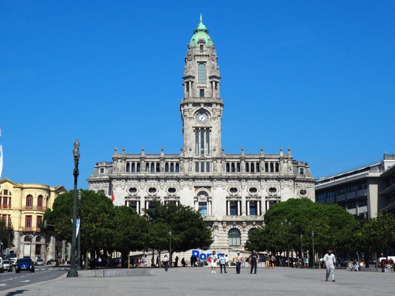 Large historic building with a clock tower, people walking in a sunny square, clear blue sky above—a perfect scene to experience during one day in Porto.