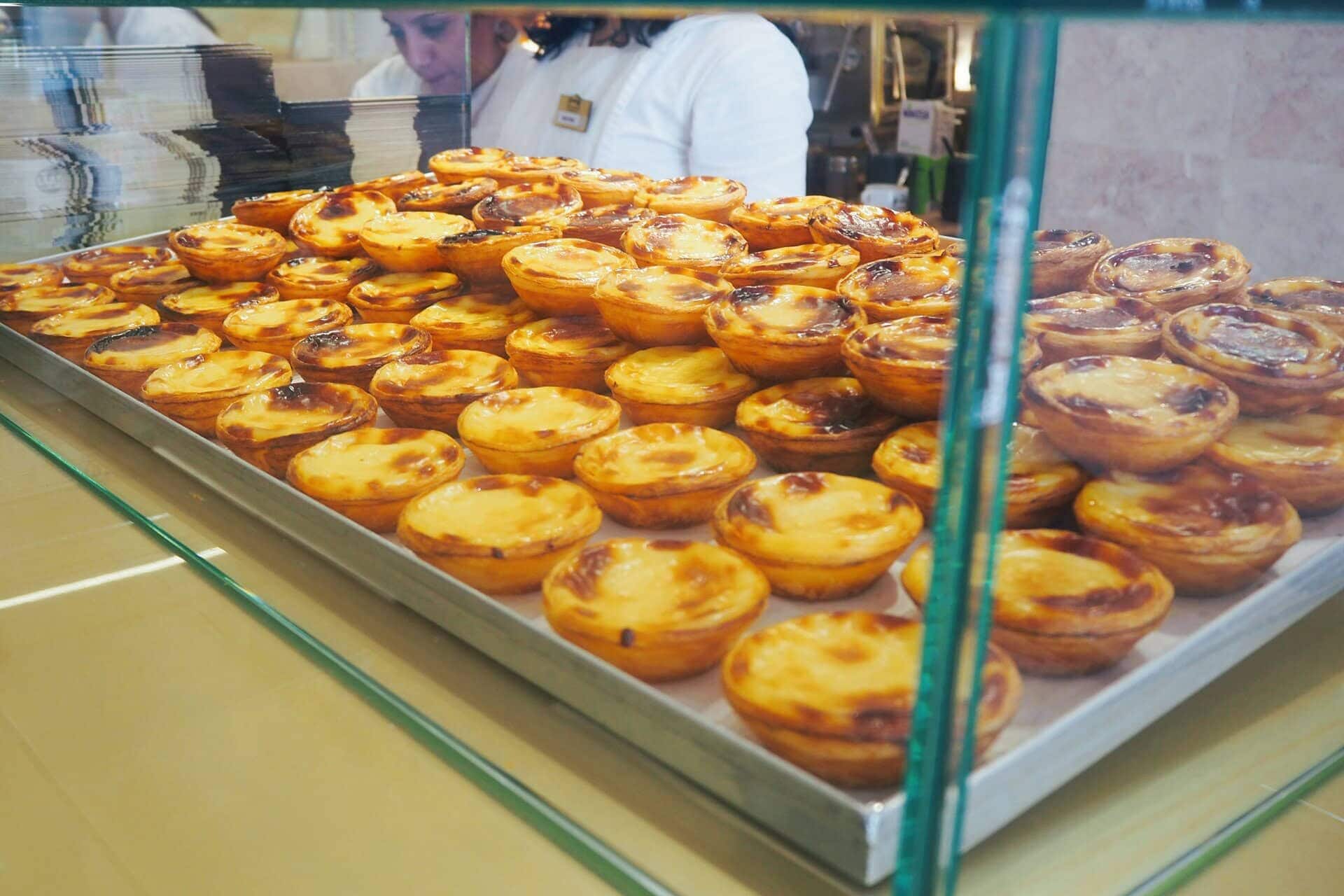 A tray filled with many custard tarts on display behind glass at a bakery counter, just waiting to be savoured by anyone spending one day in Porto.