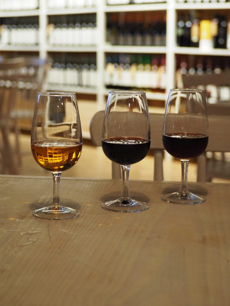 Three wine glasses—one with white wine, two with red—sit on a wooden table in front of wine shelves, evoking the rich experience of one day in Porto.