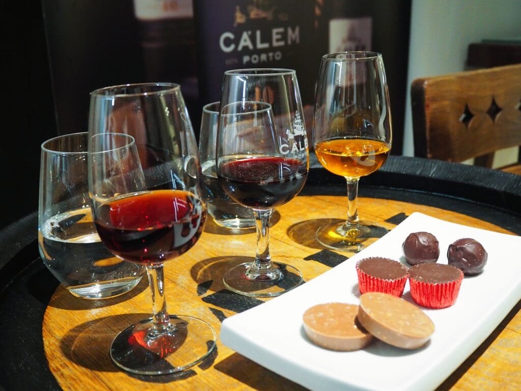 Four glasses of wine with chocolates on a white plate, set on a wooden table—an experience reminiscent of savouring the flavours you might discover during one day in Porto.