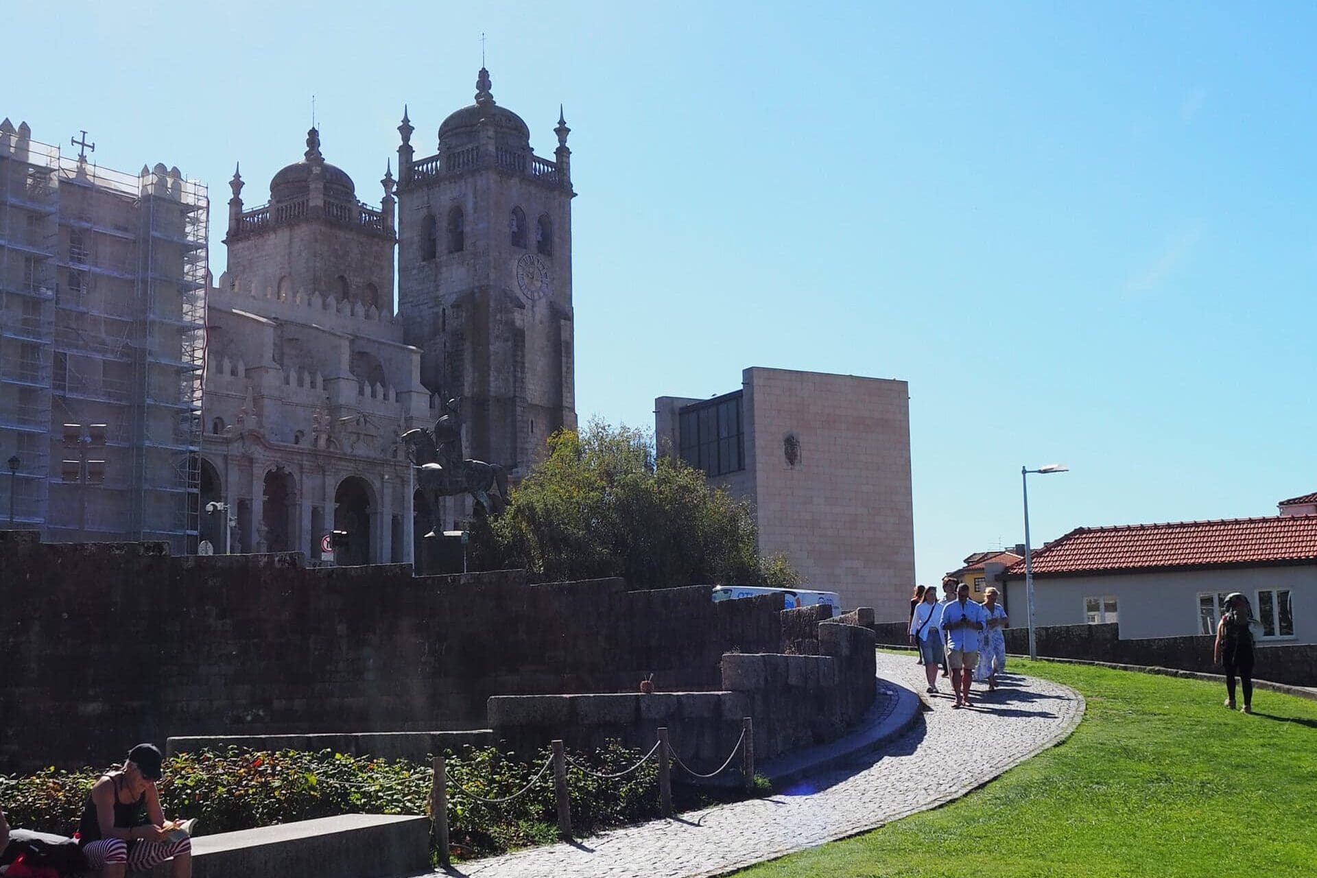 People walk on a cobblestone path near an old cathedral with towers, under a clear blue sky, soaking in the charm of one day in Porto.