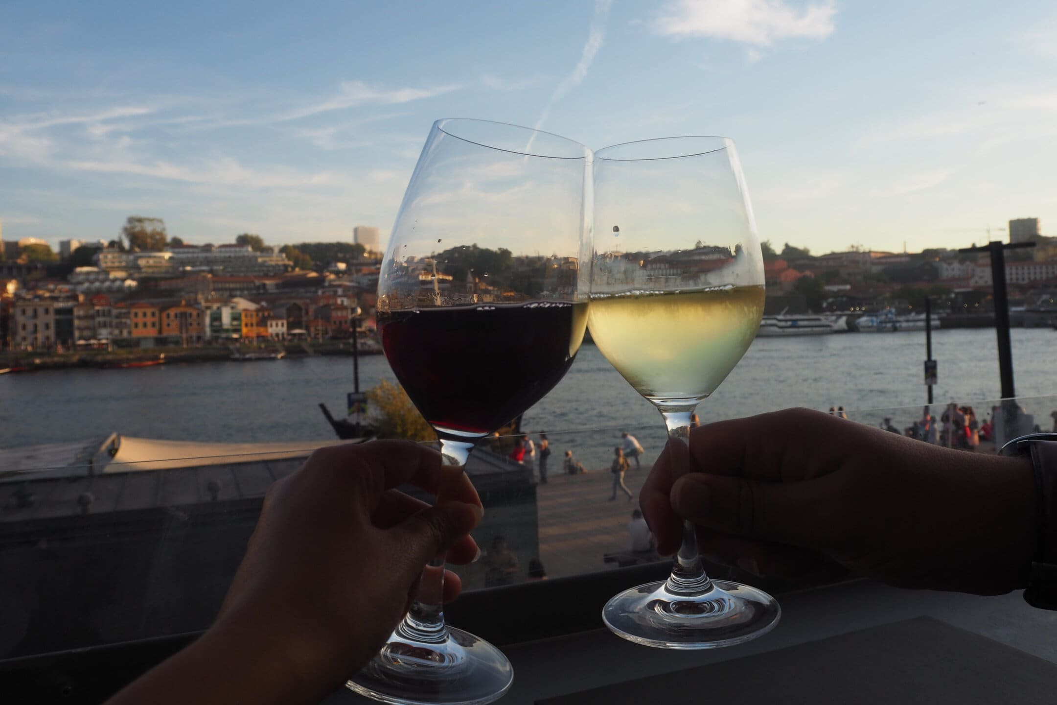 Two people clink glasses of red and white wine by a riverside city at sunset, savouring the moment in true Tips Porto style.