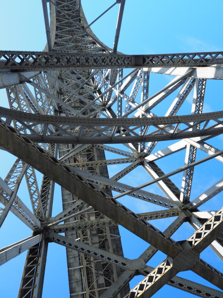 Steel structure of a tower viewed from below against a clear blue sky, reminiscent of the impressive ironwork found on the Tips Porto bridge.
