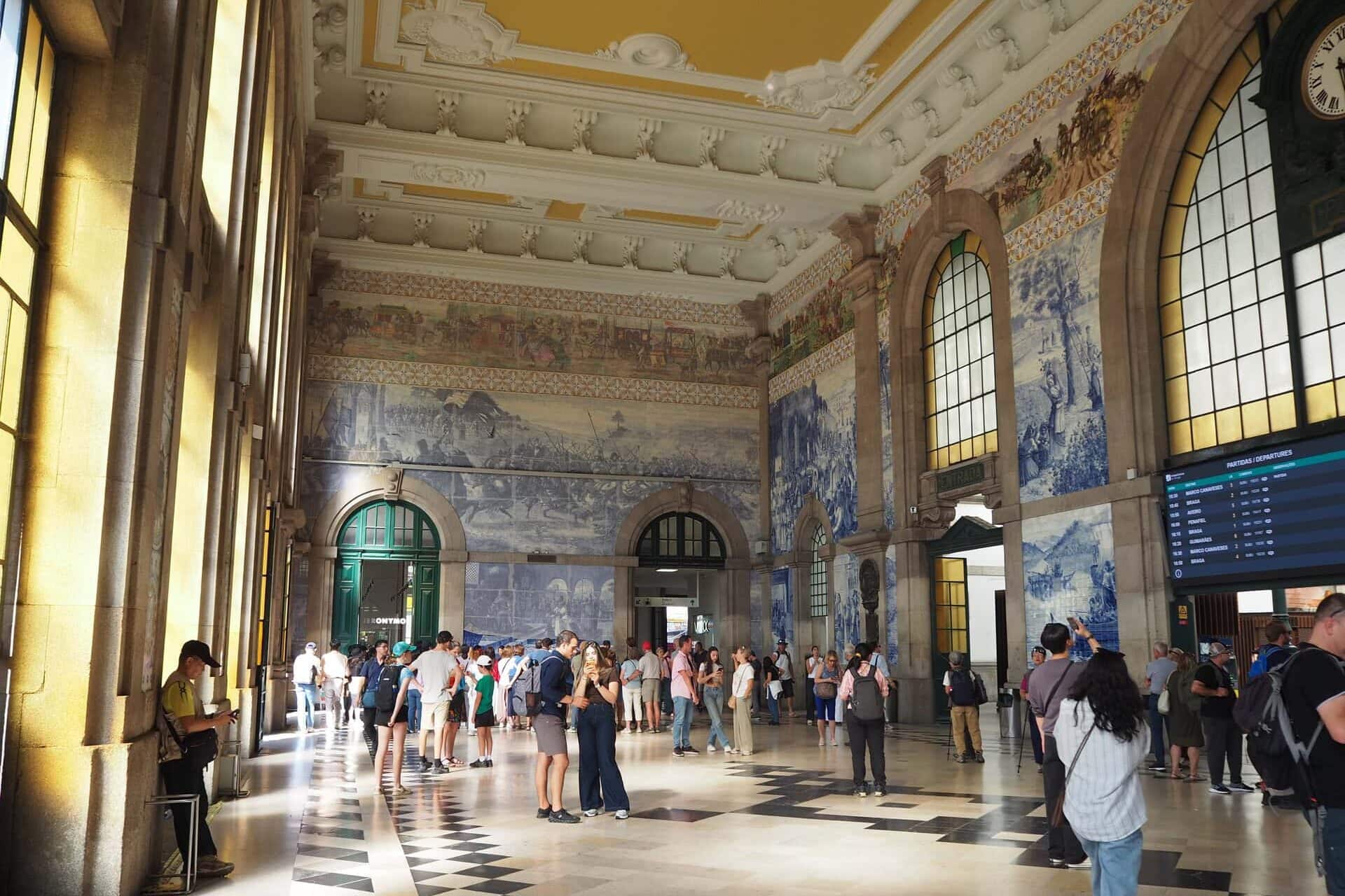 People stand in a grand railway station hall with blue tile murals, large windows, and a high ornate ceiling—an essential stop for anyone spending one day in Porto.